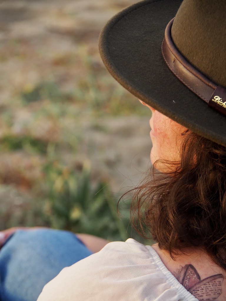 Woman will curly brown hair and a felt hat sits on the grass in demin and a white top. A tattoo is visible with a book shaped like a butterfly.