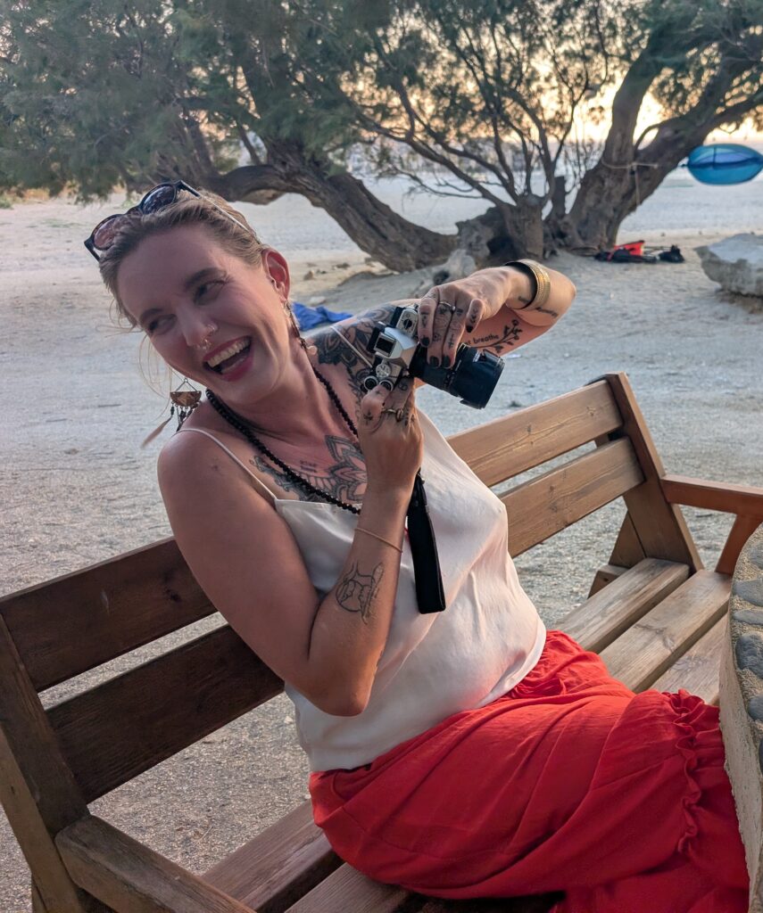 A woman in a white top and pink skirt is holding a camera and laughing. She is sitting on a bench with the beach and a tree in the background.